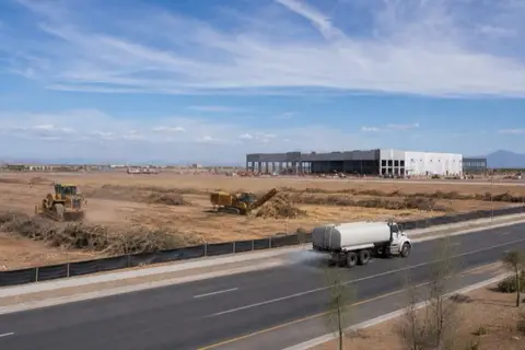 A pair of unbranded machines working a large commercial clearing along a wide Buckeye arterial with windrowed brush and a water truck controlling dust